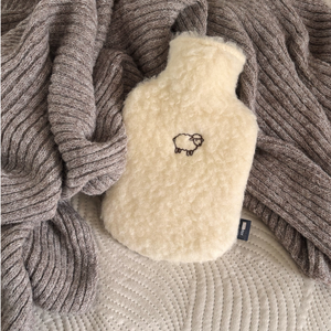 Auf einer Strickdecke platzierte Wärmflasche mit Bezug aus Bio-Wollplüsch und liebevollem Schafmotiv